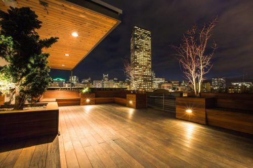 Rooftop terrace at night with city skyline, wooden flooring, and illuminated planters.