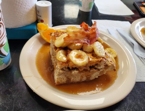 A slice of banana bread topped with bananas, bacon, and syrup, served on a plate with an orange slice.