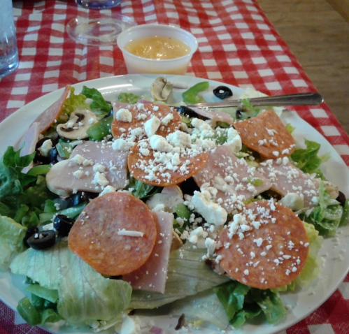 A salad with lettuce, olives, mushrooms, turkey, pepperoni, and feta cheese, served with a side of dressing.