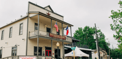 Historic building with American and Texas flags, featuring a sign for Wunsche Bros, surrounded by greenery.