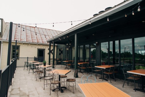 Outdoor dining area with wooden tables and chairs, string lights, and a view of a building with a metal roof.