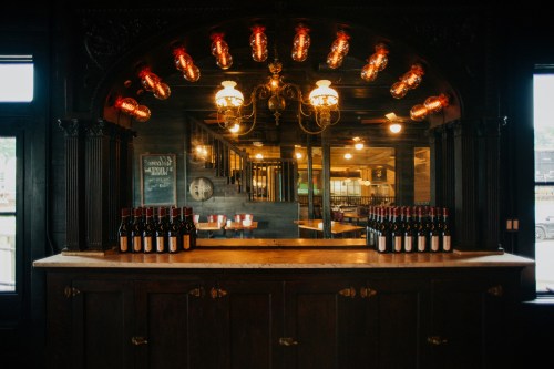 A dark wooden bar with bottles lined up, illuminated by vintage-style lights and a mirror in the background.
