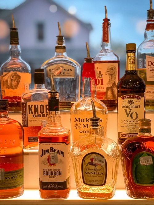 A variety of whiskey and bourbon bottles displayed on a shelf, with warm lighting and a blurred background.