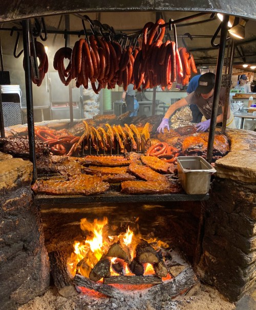 A barbecue pit with various meats cooking over an open flame, while a person prepares food in the background.