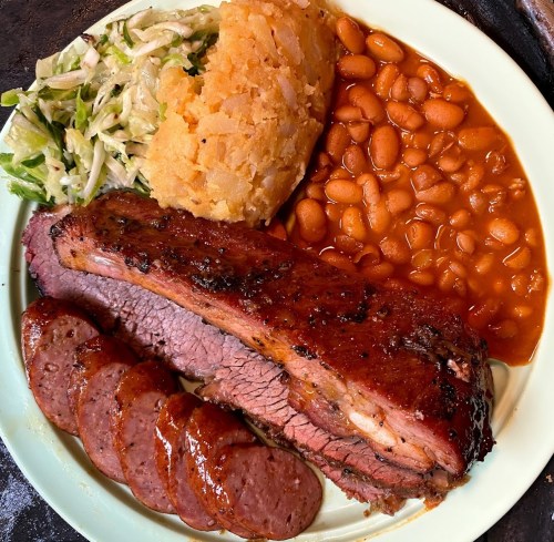 A plate of barbecue featuring sliced brisket, sausage, baked beans, coleslaw, and hash browns.