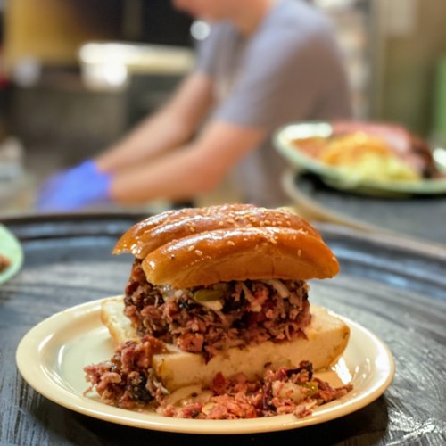 A close-up of a sandwich with shredded meat and pickles on a plate, with blurred kitchen activity in the background.