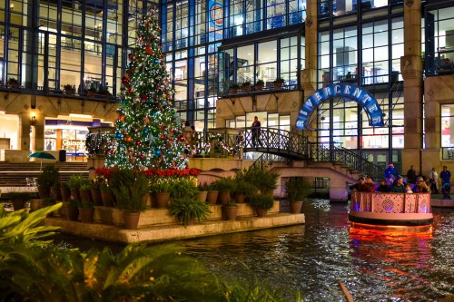A festive scene featuring a decorated Christmas tree by a waterway, with people enjoying the ambiance in a shopping center.