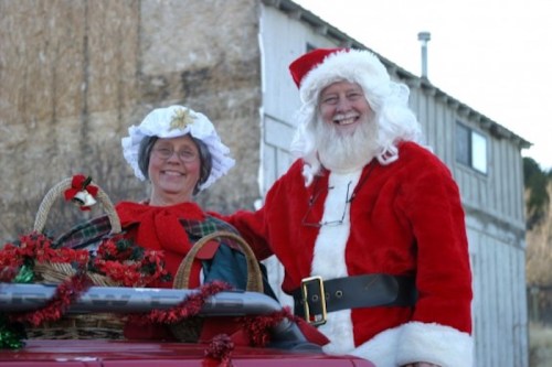 A smiling Santa Claus and a woman in festive attire pose together beside a decorated vehicle.