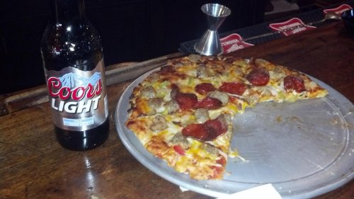 A slice of pizza with toppings next to a bottle of Coors Light on a bar counter.