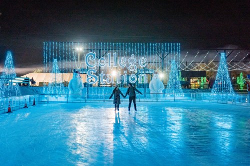 A couple ice skates on a rink illuminated by blue lights, with "College Station" sign and festive decorations in the background.