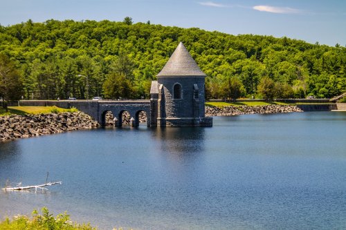 A stone tower with a conical roof stands by a calm lake, surrounded by lush green trees and a clear blue sky.