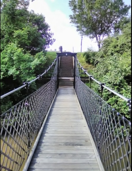 A wooden suspension bridge leads to a grassy hill, surrounded by lush greenery and a clear blue sky.