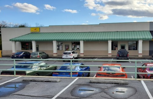 A row of parked cars in front of a commercial building with a green roof and a sign. Cloudy sky in the background.