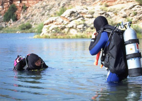 Two divers in wetsuits are partially submerged in a lake, preparing for a dive amidst rocky surroundings.