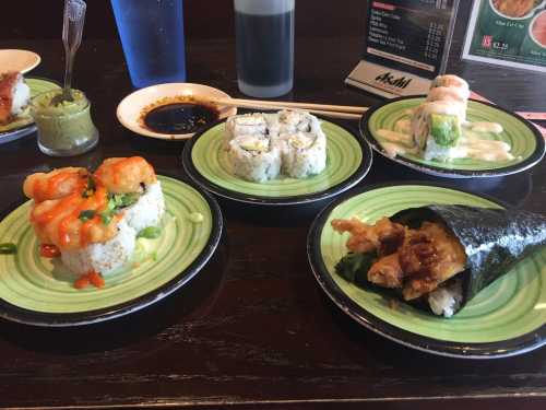 A variety of sushi dishes on green plates, including shrimp rolls, California rolls, and a hand roll, with soy sauce and wasabi.