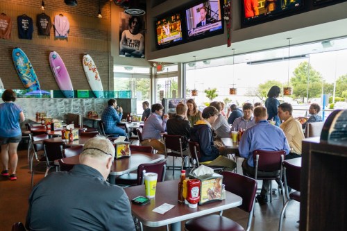 A busy restaurant scene with diners at tables, a TV showing news, and surfboards decor on the walls.