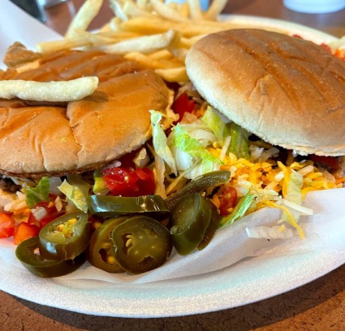 Two burgers with lettuce, tomatoes, cheese, jalapeños, and a side of French fries on a white plate.