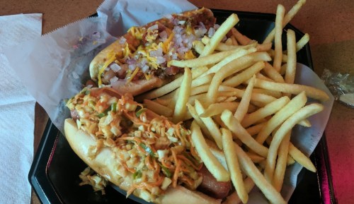 Two hot dogs topped with onions and chili, served with a side of crispy French fries on a black tray.
