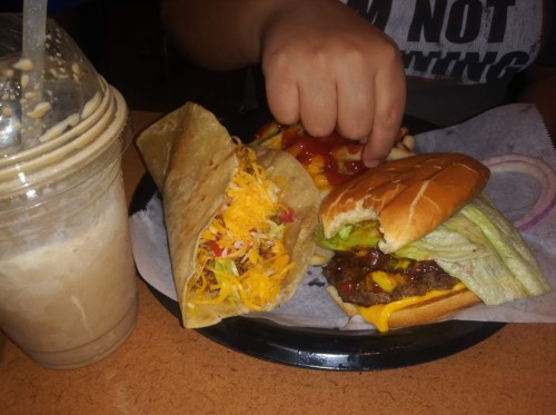 A hand reaching for a burger and taco on a plate, with a milkshake on the side.