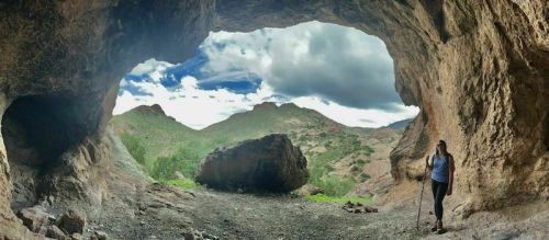 A hiker stands inside a cave, looking out at a scenic landscape of mountains and cloudy skies.