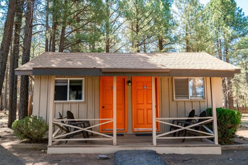 A small cabin in a forest, featuring two orange doors and a porch with chairs, surrounded by tall pine trees.