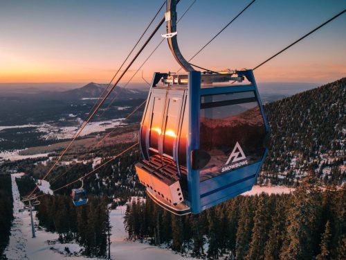 A blue gondola lifts over snowy terrain at sunset, surrounded by mountains and trees.