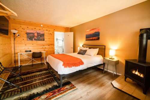 Cozy hotel room with a wooden interior, a queen bed, a small table, and a fireplace, featuring warm lighting and decor.