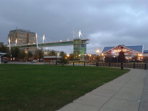 A modern pedestrian bridge illuminated at dusk, connecting buildings over a grassy area with a nearby entertainment venue.