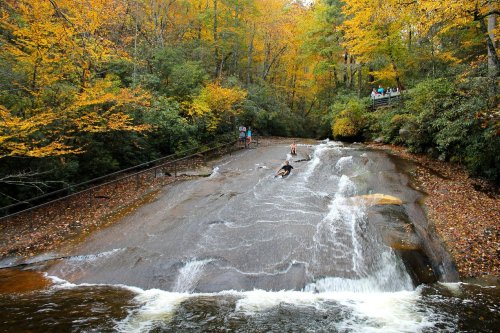 A scenic waterfall flows over smooth rocks, surrounded by vibrant autumn foliage and people enjoying the view.