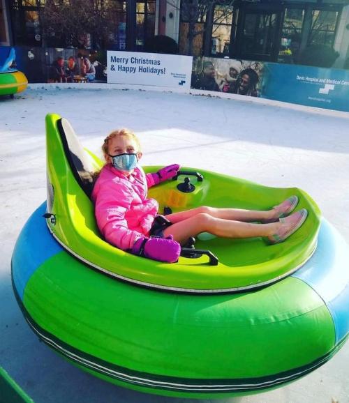 A girl in a pink jacket and mask sits in a green inflatable bumper car on an ice rink, smiling at the camera.