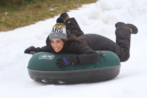A woman in winter gear joyfully sleds down a snowy hill on a green inflatable tube.