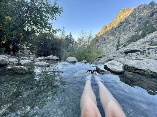 A person’s legs in sandals relax in a clear, shallow stream surrounded by rocky terrain and trees.