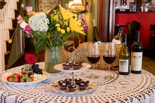 A table set with a lace tablecloth, featuring flowers, wine bottles, glasses, fruit, cheese, and chocolate cupcakes.