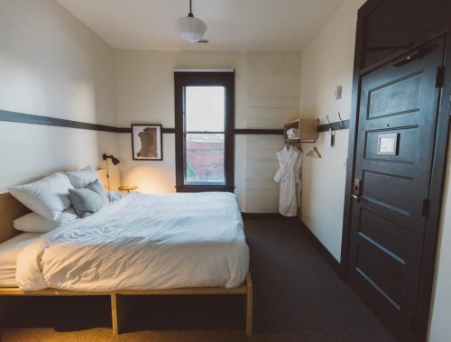 A cozy hotel room featuring a bed, window, and a robe hanging on a hook. Minimalist decor with warm lighting.