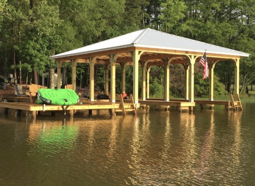A wooden dock with a covered area, surrounded by water and trees, featuring an American flag and outdoor seating.
