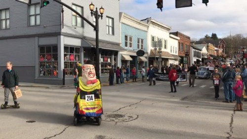 A colorful clown figure on a small cart parades down a street lined with spectators and shops.