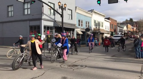 A festive parade with people on bicycles, some in costumes, riding down a street lined with shops and spectators.