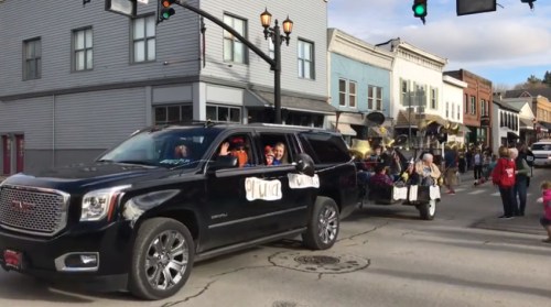 A black SUV with passengers waves during a parade, followed by a trailer decorated with festive items.