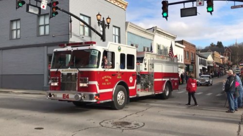 A red fire truck drives through a small town intersection, with buildings and pedestrians in the background.