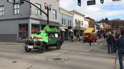 A green car decorated like a dragon leads a parade down a street lined with shops and spectators.