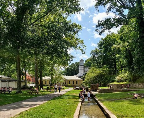 A sunny park scene with people walking along a path, a stream, and lush green trees, featuring a building in the background.