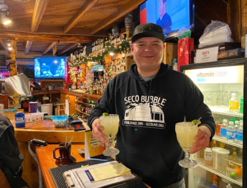 A smiling bartender holds two cocktails at a lively bar, with colorful decorations and a TV in the background.