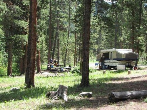 A campsite surrounded by tall trees, featuring a trailer and a picnic area with people gathered around a table.