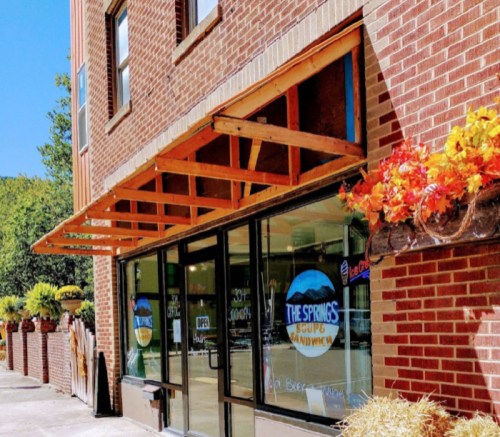 Exterior of a sandwich shop with large windows, wooden awning, and colorful flower pots. Sign reads "The Springs Soup & Sandwich."