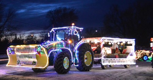 A brightly lit tractor with festive decorations and a trailer, set against a twilight sky.