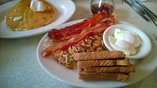 A plate of breakfast featuring bacon, hash browns, poached eggs, toast, and a side of pancakes with butter.