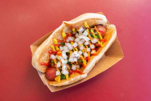 A hot dog in a bun topped with diced onions, mustard, ketchup, and relish, served in a cardboard tray.