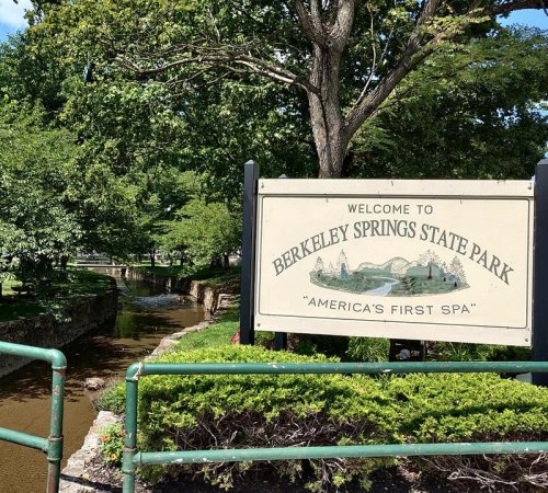 Sign welcoming visitors to Berkeley Springs State Park, featuring lush greenery and a stream nearby.