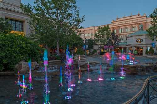 Colorful water fountains illuminated at dusk, surrounded by greenery and buildings in a lively outdoor setting.