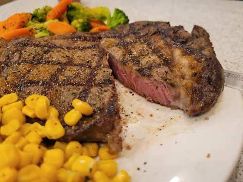 A grilled steak with a juicy pink center, served with corn and a side of mixed vegetables.